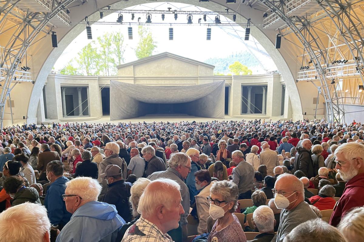 A view of the audience in the midst of the theater at Oberammergau shows hundreds of older, white haired attendees, some wearing face masks, taking their seats under an arched canopy.