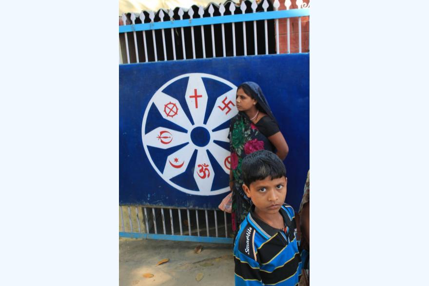 A boy in a blue striped shirt and a woman in a sari in headscarf stand before the blue gate to Matridham Ashram which displays symbols of the major religious traditions of the Indian subcontinent.