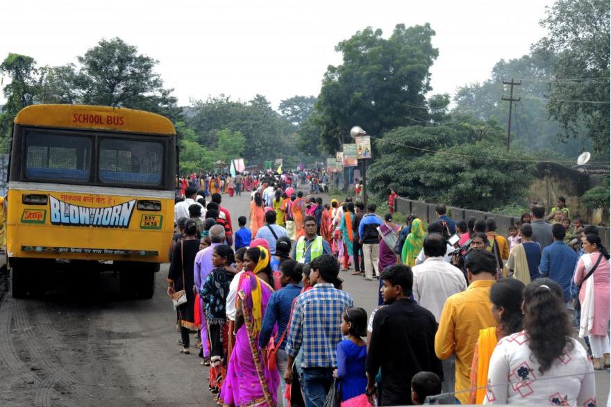 Lines of devotees in procession along a rural road, walk around a yellow bus.