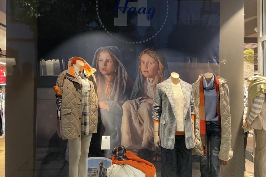 The window display of a local store combines mannequins wearing current fashions with large billboards of young girls dressed in their Passion Play costumes.