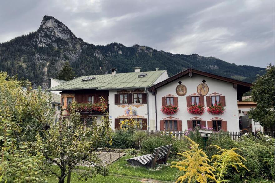 A building's exterior adorned with decorative painting and religious scenes, with a small garden area in front and the alps behind.
