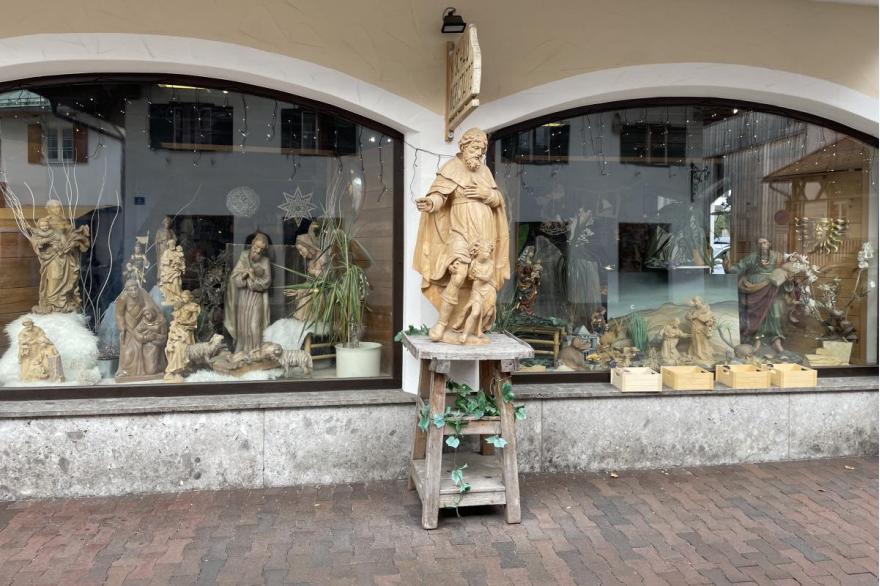 A storefront for a woodcarving business with an elaborate sculpture on a wooden pedestal draped with ivy out front .