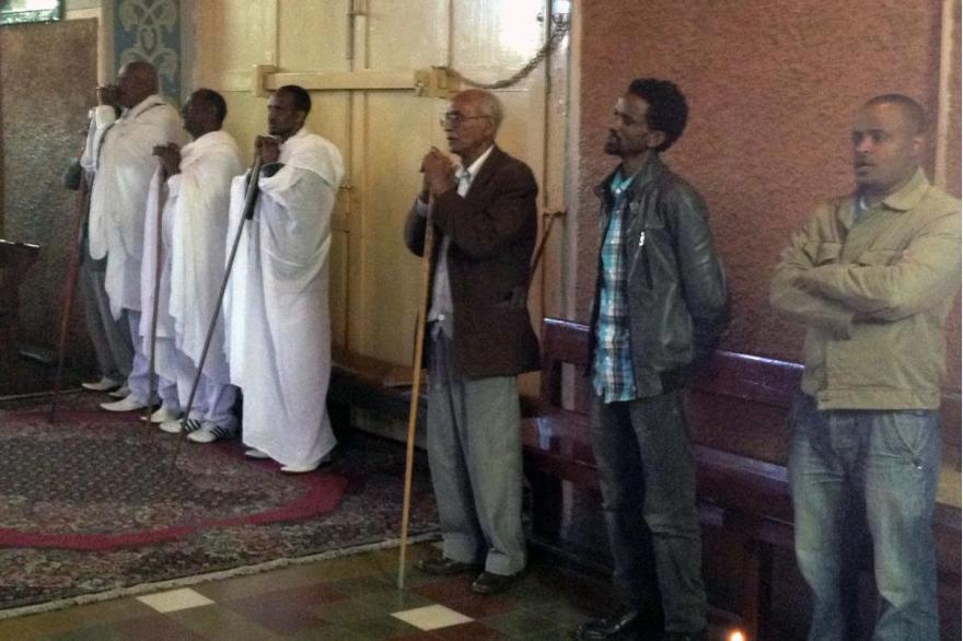 Men pray at the back of Mass in the traditional Ethiopian fashion by standing with the aid of a prayer staff throughout the liturgy. Staffs are kept int he back of the church for those who need them.