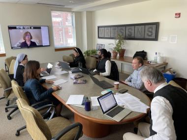 Symposium participants sit around a conference table before a large screen where Prof. Wanner and international participants join by Zoom.
